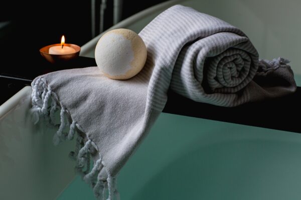 "A serene spa-like bathroom scene featuring a High Hopes Co CBD Bath Bomb infused with 100mg CBD isolate. The bath bomb rests on a neatly rolled striped towel beside a lit candle in a copper holder, creating a warm, relaxing atmosphere."